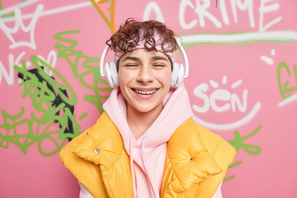 Positive curly haired youngster smiles gladfully has braces on teeth dressed in hoodie with vest enj